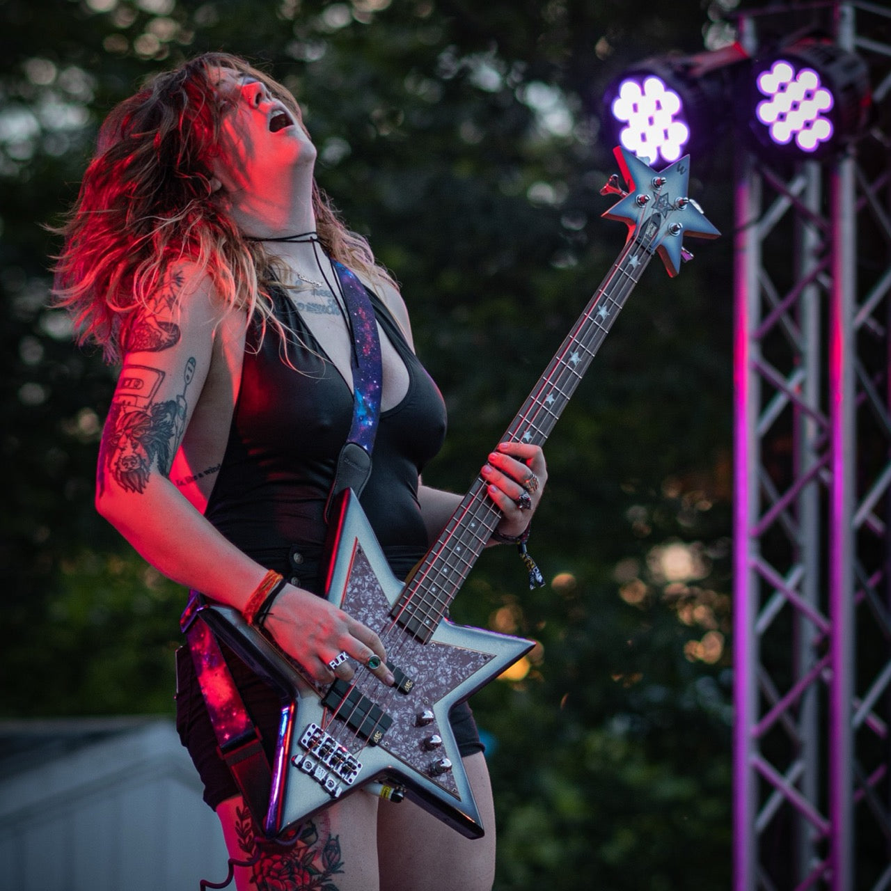 Image shows a person playing guitar on stage. They are dressed in leather  with long hair and tattoos. The guitar is star shaped. 