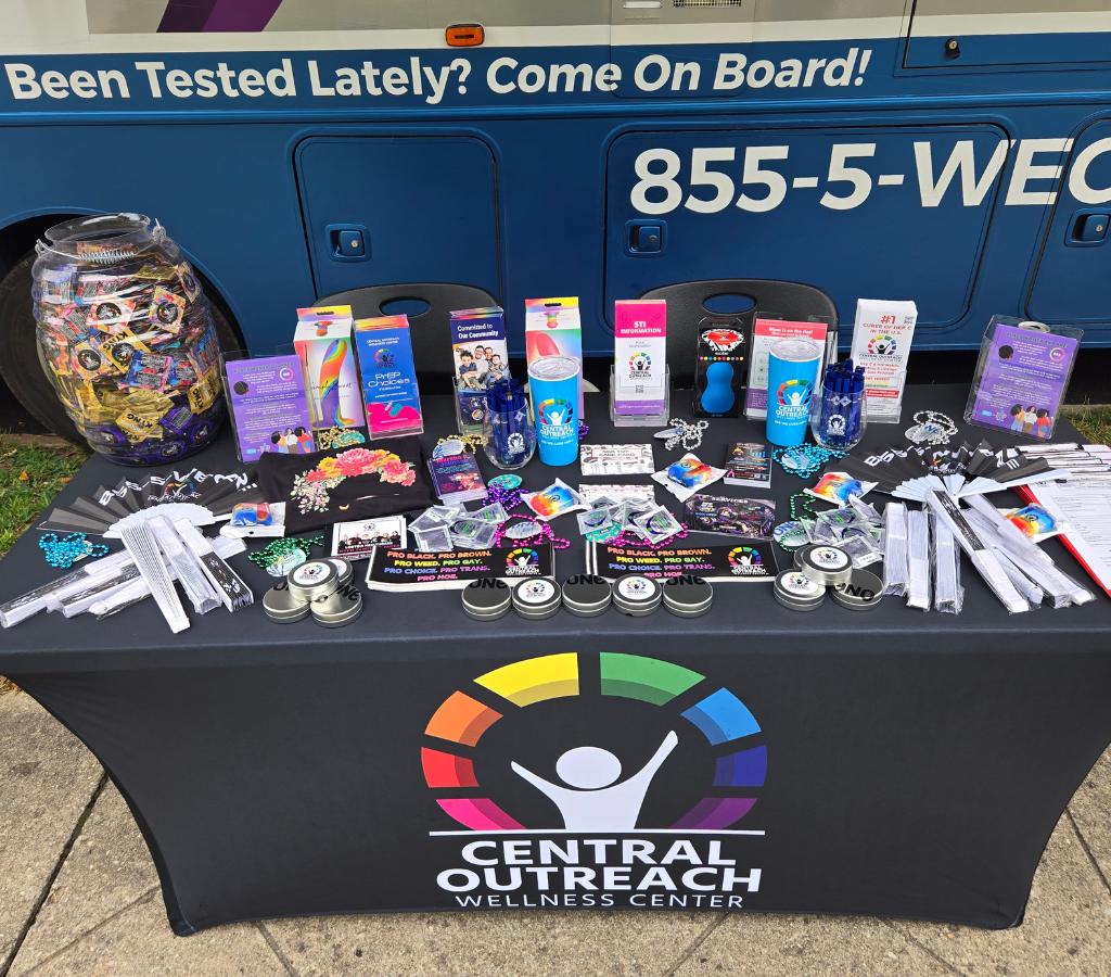 A table at an event outside. The table has the Central Outreach logo on the table cloth, and lots of condoms, water bottles, educational materials, and more on the table.
